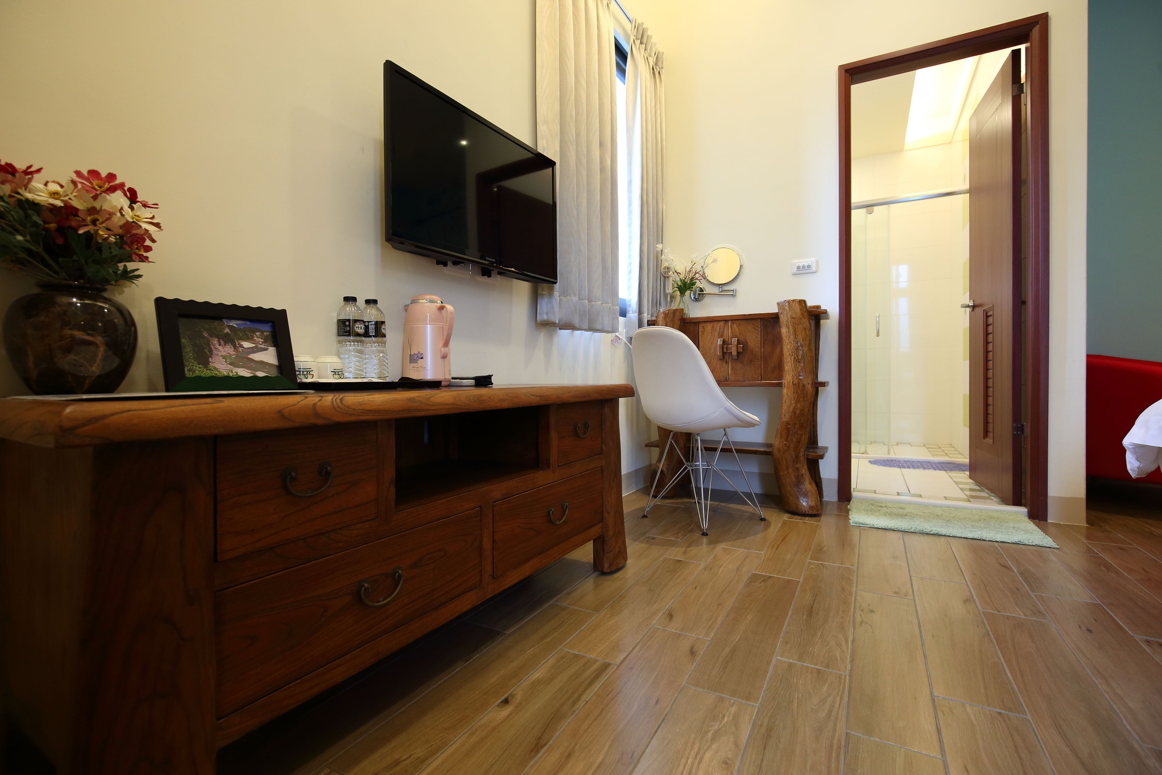 A wooden dresser with drawers, a flat-screen TV mounted on the wall, a white chair, a small table with a vase of flowers, and a door leading to another room.
