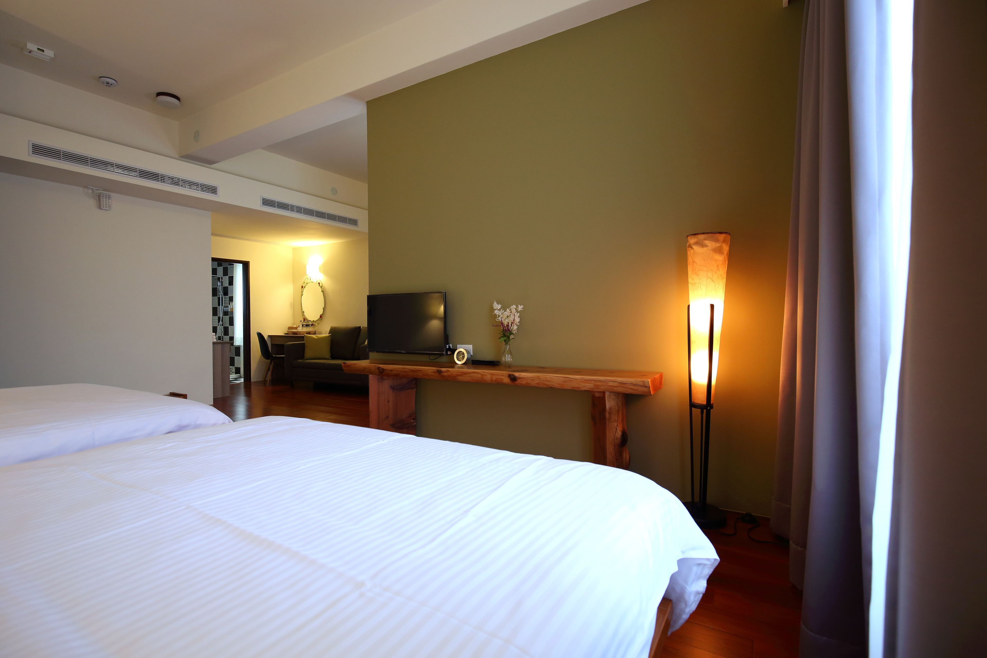Una habitación de hotel con una cama grande, un escritorio con televisor, una lámpara y un pequeño jarrón con flores.
