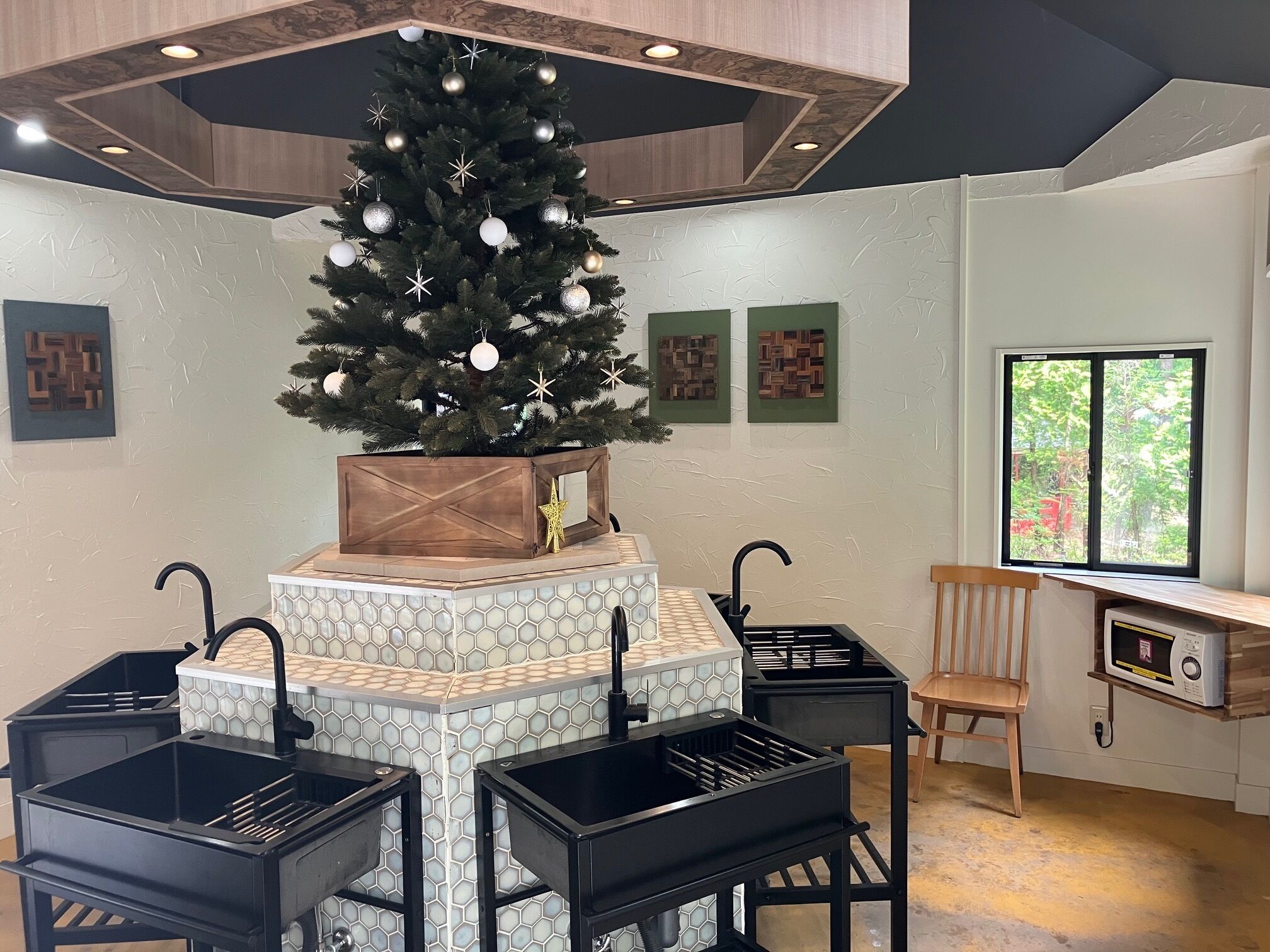 A modern kitchen with a central island, black bar stools, a wooden chair, a Christmas tree, and a window with a view.