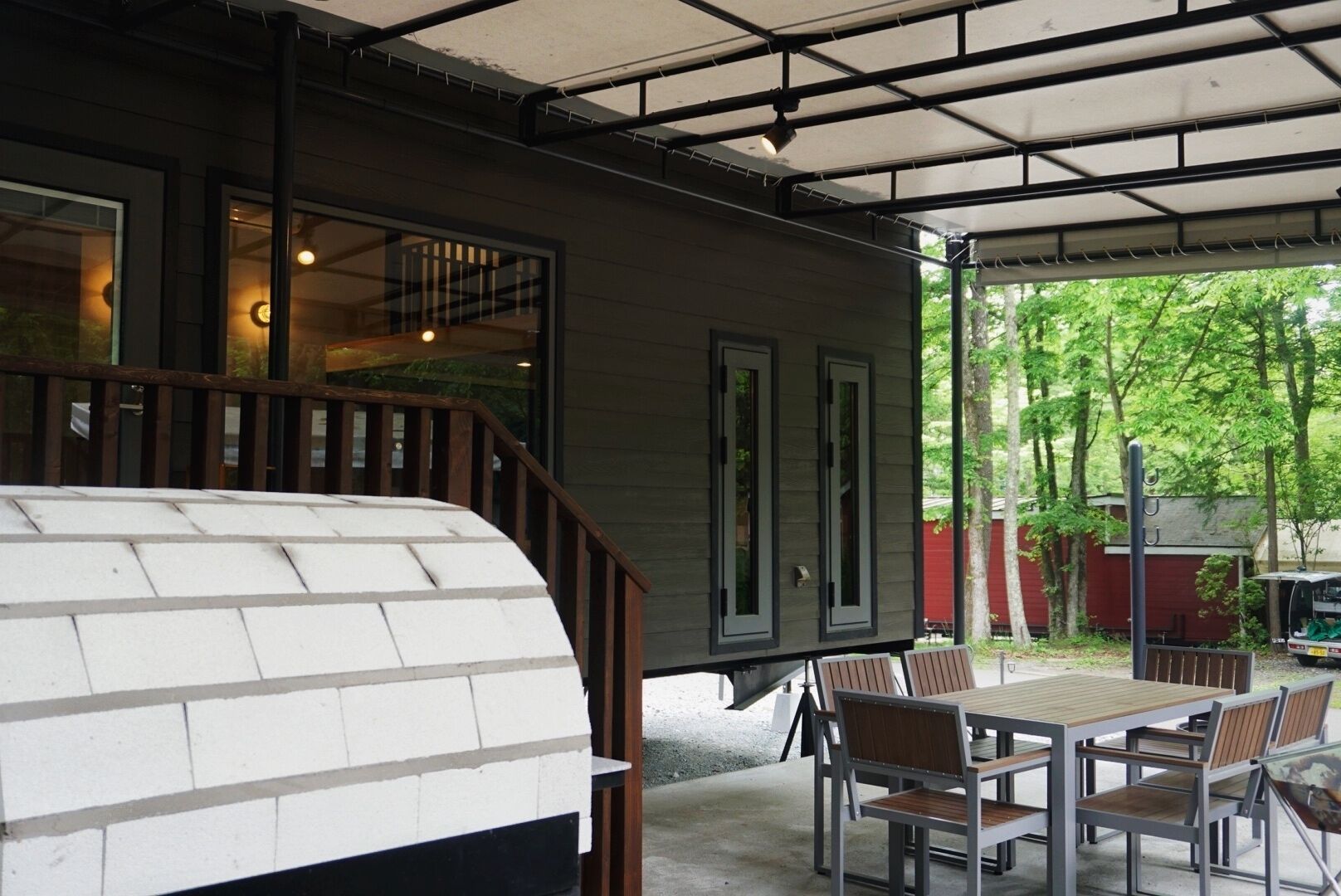 A covered patio area with a table and chairs, a staircase leading to an upper level, and a view of trees and a red building in the background.