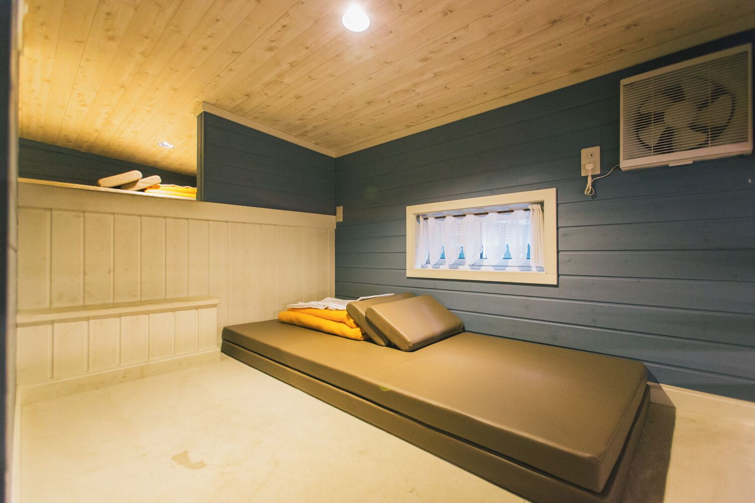 A small, single-bed room with a wooden ceiling, a window with curtains, and a wall-mounted fan.