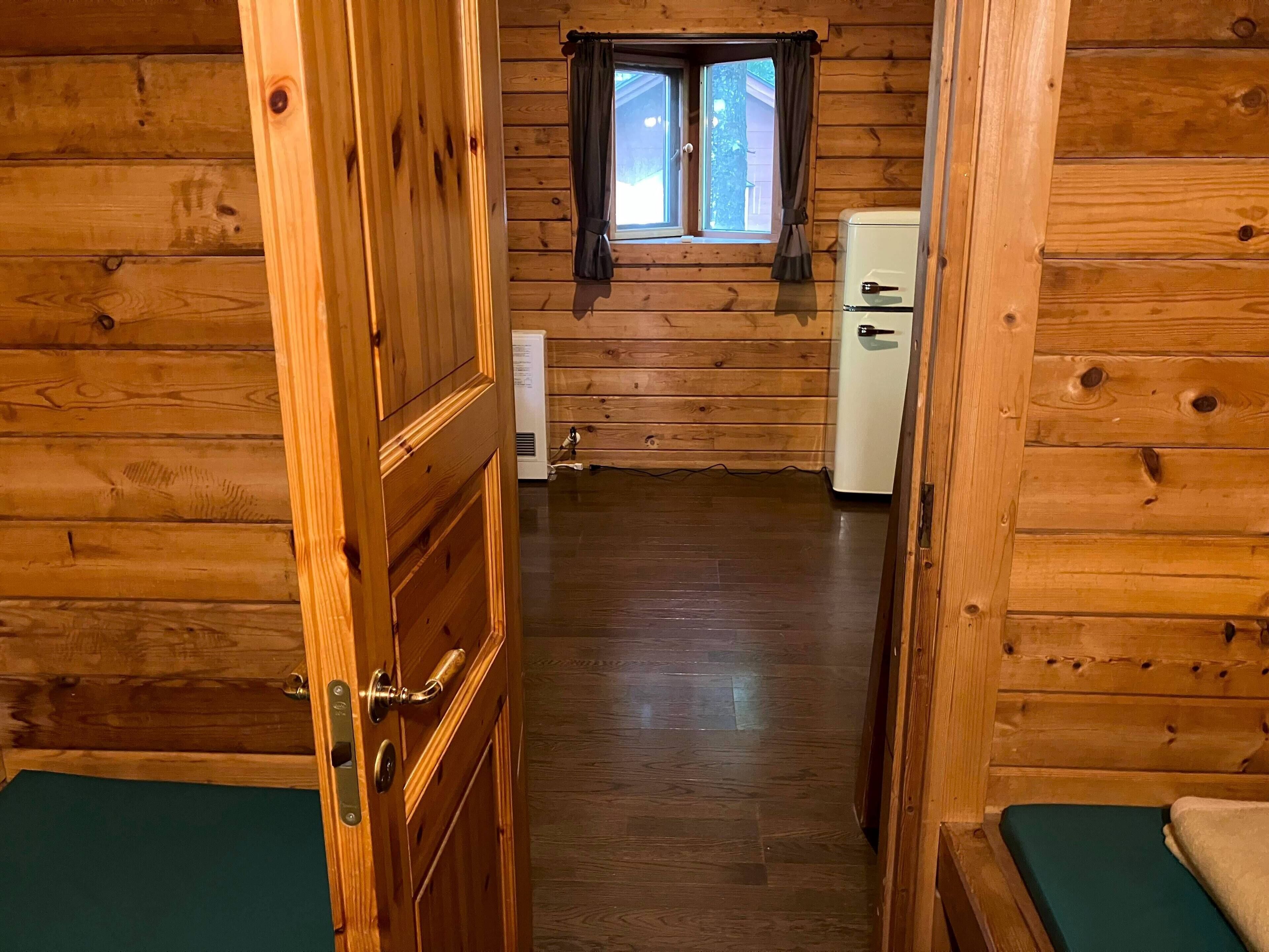 Interior de una cabaña de madera con puerta de madera, ventana con cortinas, un refrigerador blanco y piso de madera.
