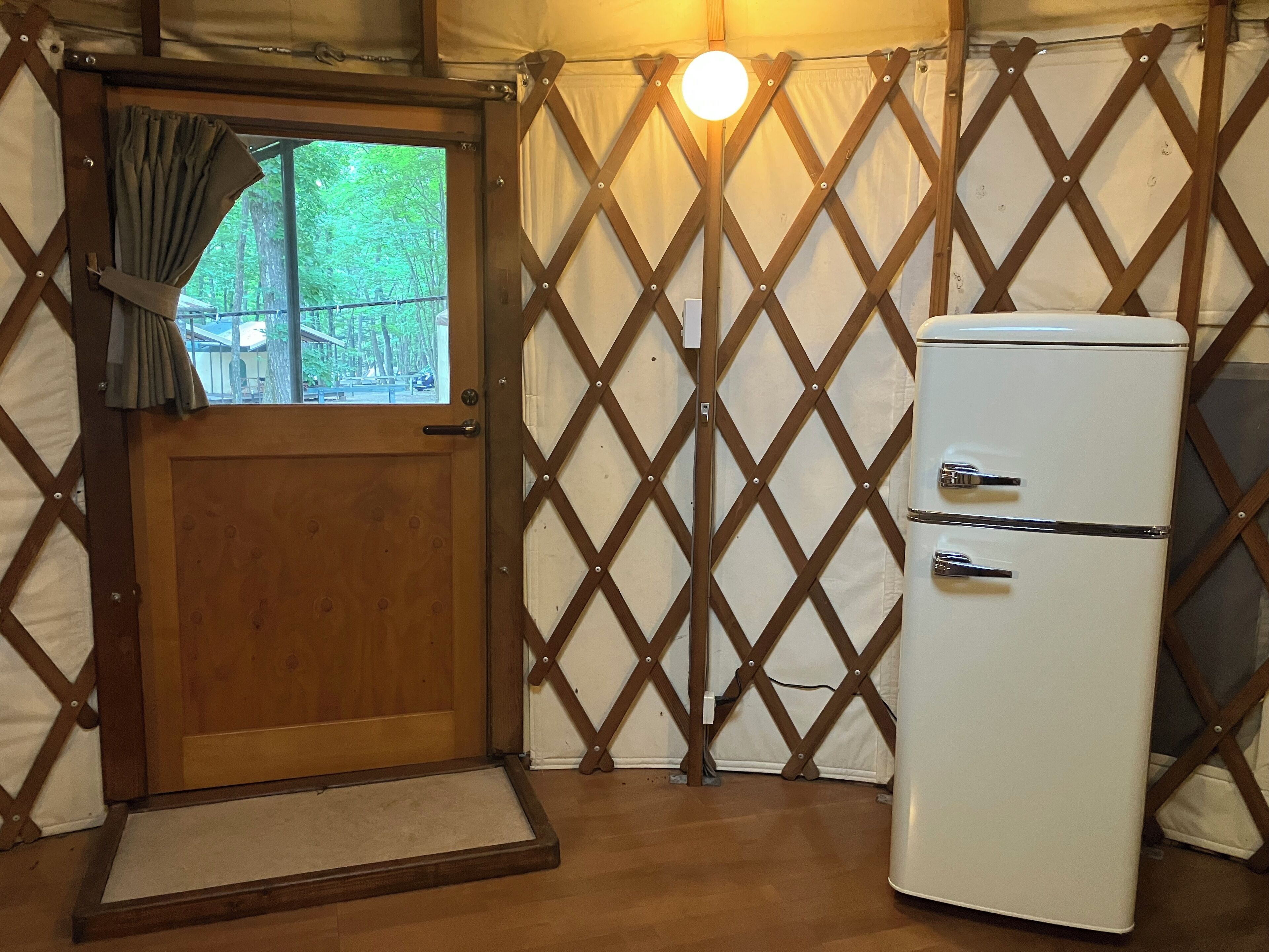 Une pièce de taille modeste avec une porte en bois, une petite fenêtre avec des rideaux, un réfrigérateur blanc et une cloison ajourée.