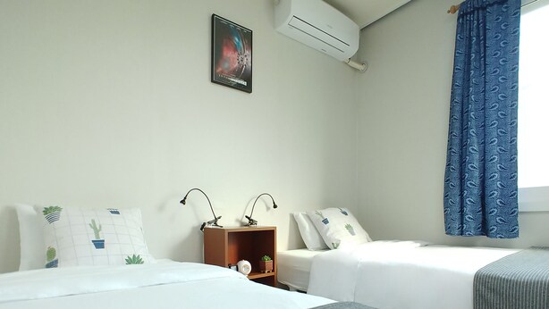 A hotel room with two beds, a nightstand, a wall-mounted air conditioner, a framed picture, and a window with blue curtains. - Hause Itaewon - Hostel (Seoul)
