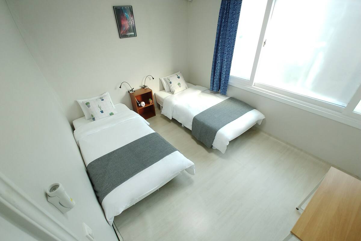 A hotel room with two beds, a small wooden nightstand, a wall-mounted picture, and a window with curtains.