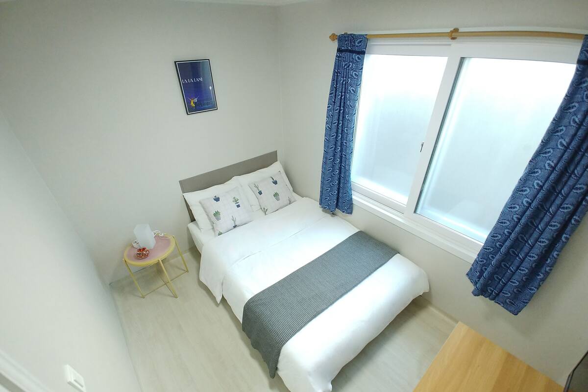 A neatly arranged bedroom with a bed, a bedside table, a small stool, and a window with curtains.