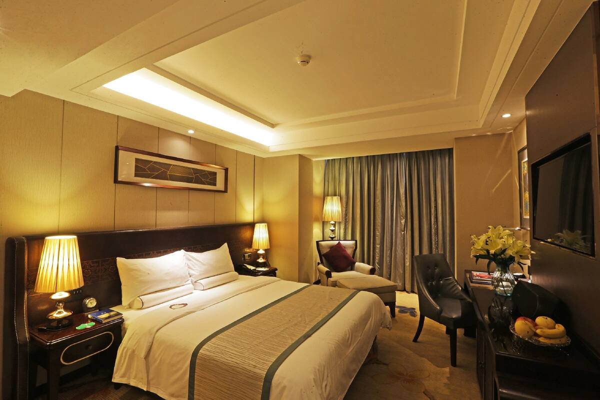 A hotel room with a large bed, two bedside tables with lamps, a chair, a desk with a vase of flowers, and a television on the wall.