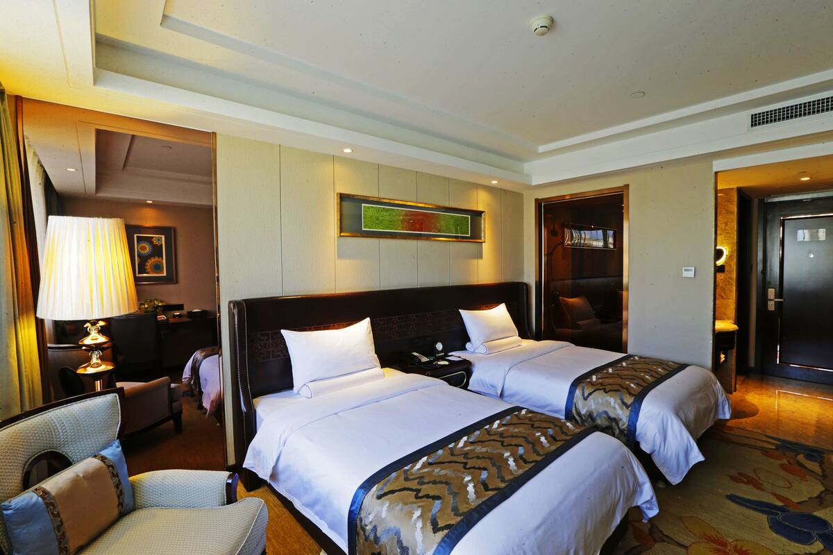 A hotel room with two beds, a desk, a chair, and a lamp.