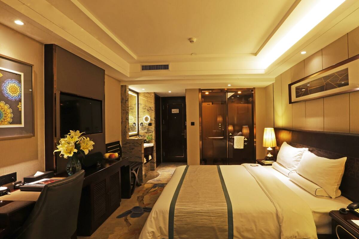 A hotel room with a bed, a desk with a computer, a TV, a mirror, and a bathroom with a shower and toilet.