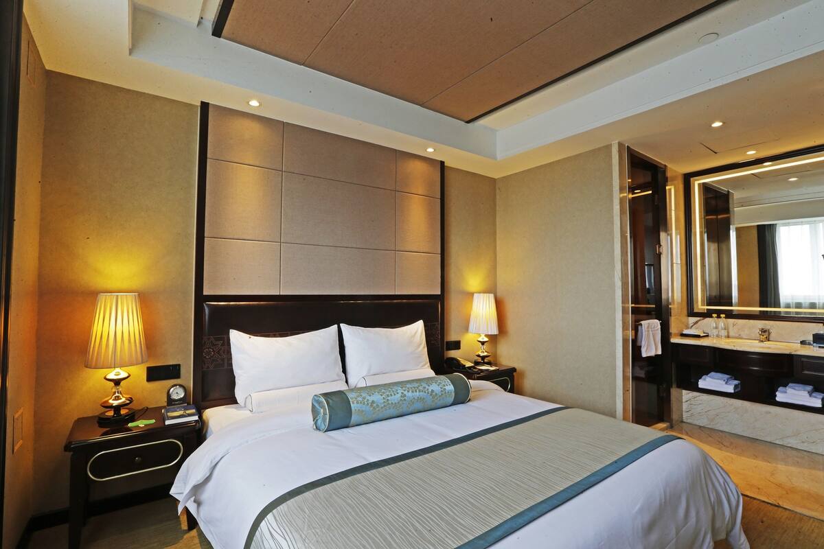 A hotel room with a large bed, two bedside tables with lamps, a headboard, a bathroom with a mirror, and a towel rack.