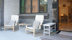 Two white rocking chairs with patterned cushions, a small round table, and a white side table with shoes on it, outside a building with a glass door and a brick wall. - Dio Inn (Hengchun)