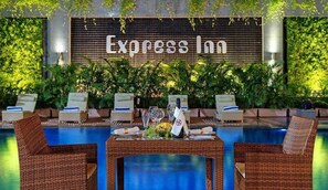 A poolside area with wicker furniture and a table set for dining, surrounded by lush greenery and a sign reading "Express Inn". - Express Inn Nashik (Nashik)