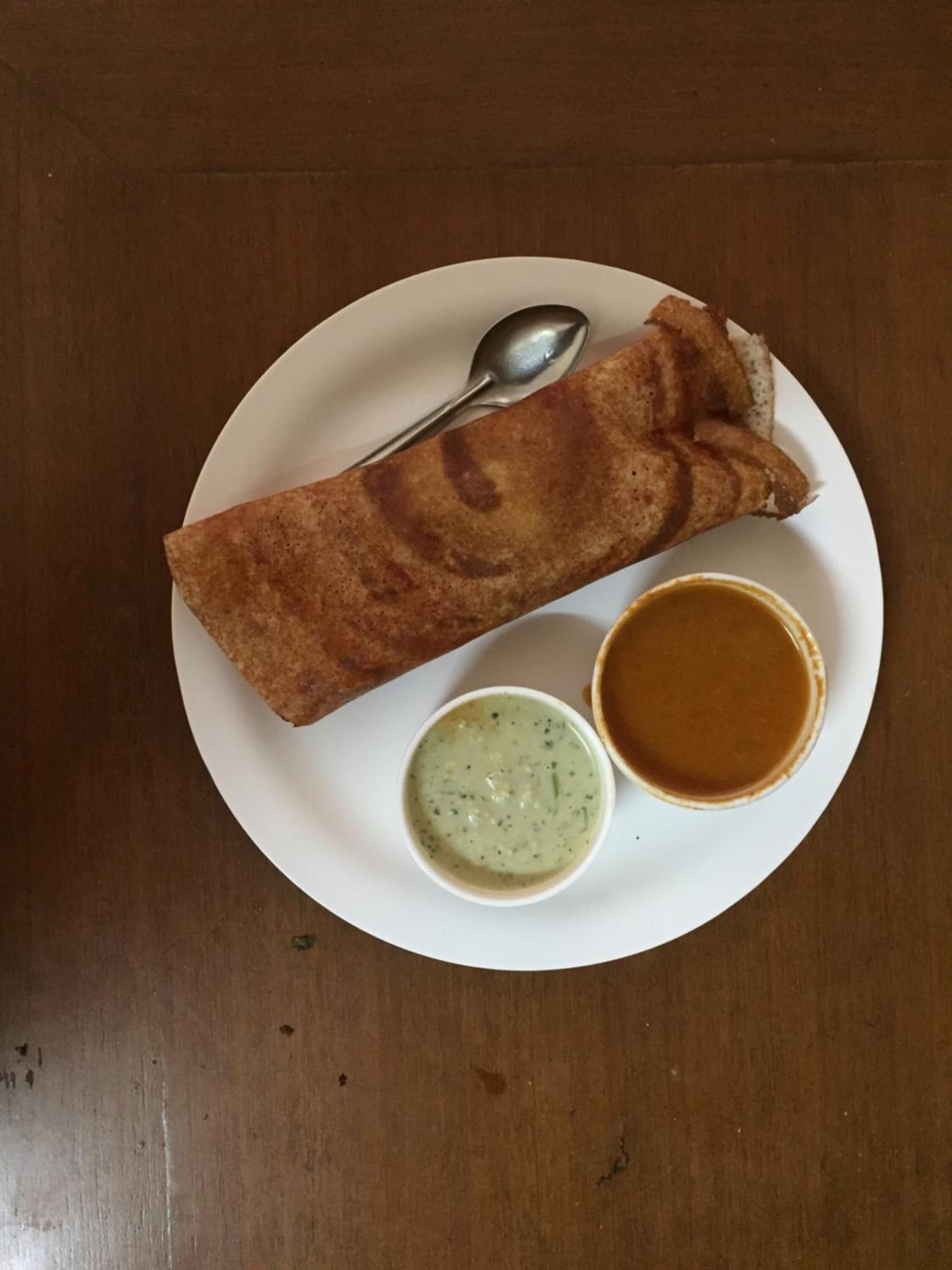 A plate with a folded crepe, two small bowls of sauce, and a spoon on a wooden table.