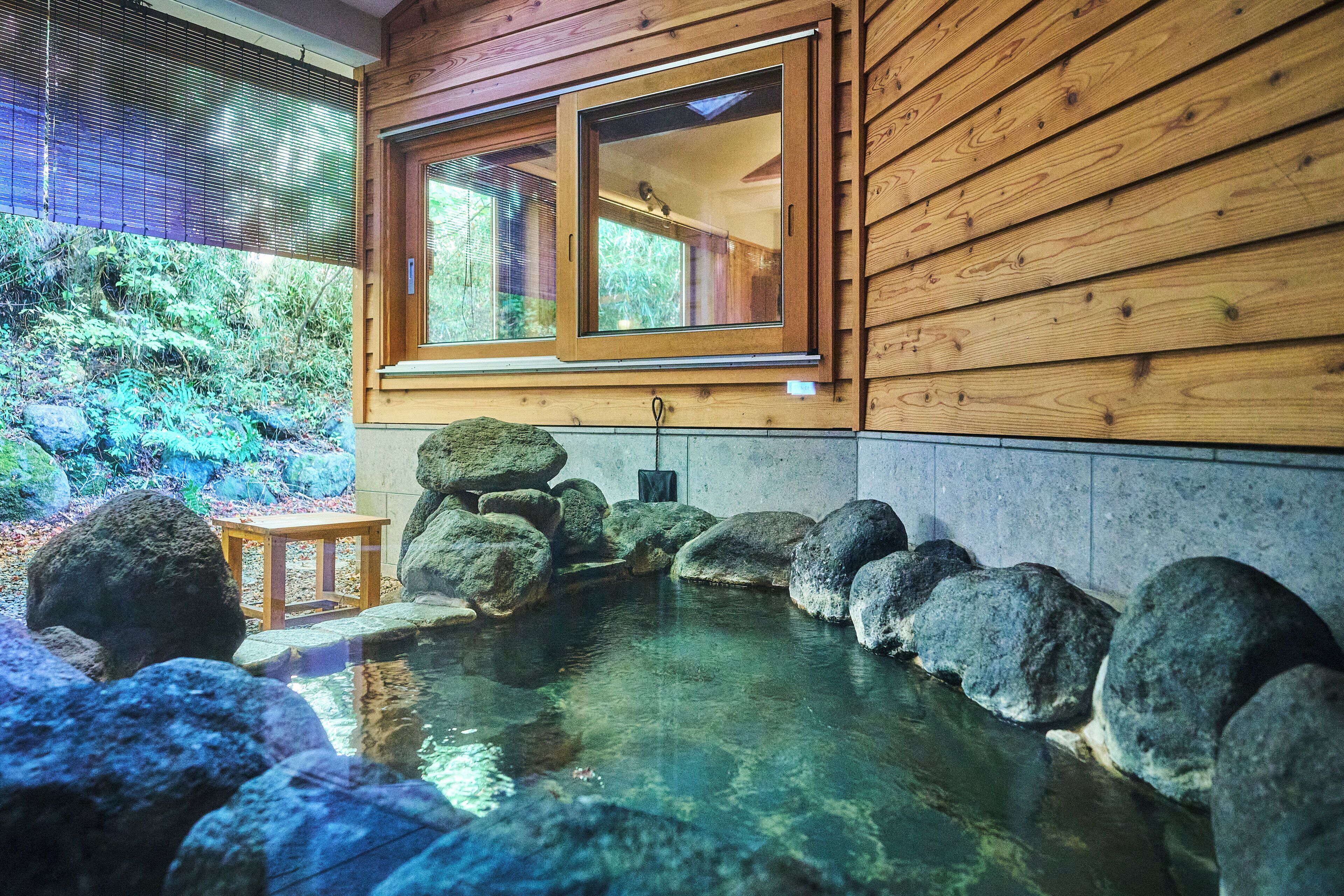 A wooden cabin with a stone-lined pond, a wooden table, and a window overlooking greenery.