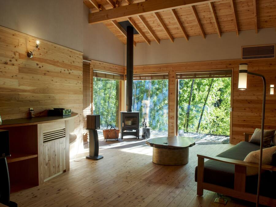 A spacious room with a wooden interior, a bed, a sofa, a fireplace, and large windows offering a view of the outdoors.