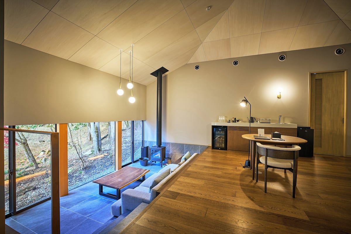 Un intérieur moderne avec un plancher en bois, un coin salon comprenant un banc et des coussins, une table à manger avec des chaises, un espace cuisine et de grandes fenêtres offrant une vue sur l’extérieur.