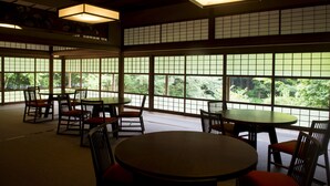 Um quarto tradicional japonês com piso de esteiras de tatame, mesinhas e cadeiras baixas. Grandes janelas com painéis shoji oferecem vista da vegetação ao redor.
