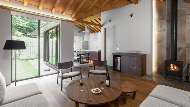 Een moderne woonkamer met een ronde houten tafel, twee banken, een open haard en een keuken met een groot raam.