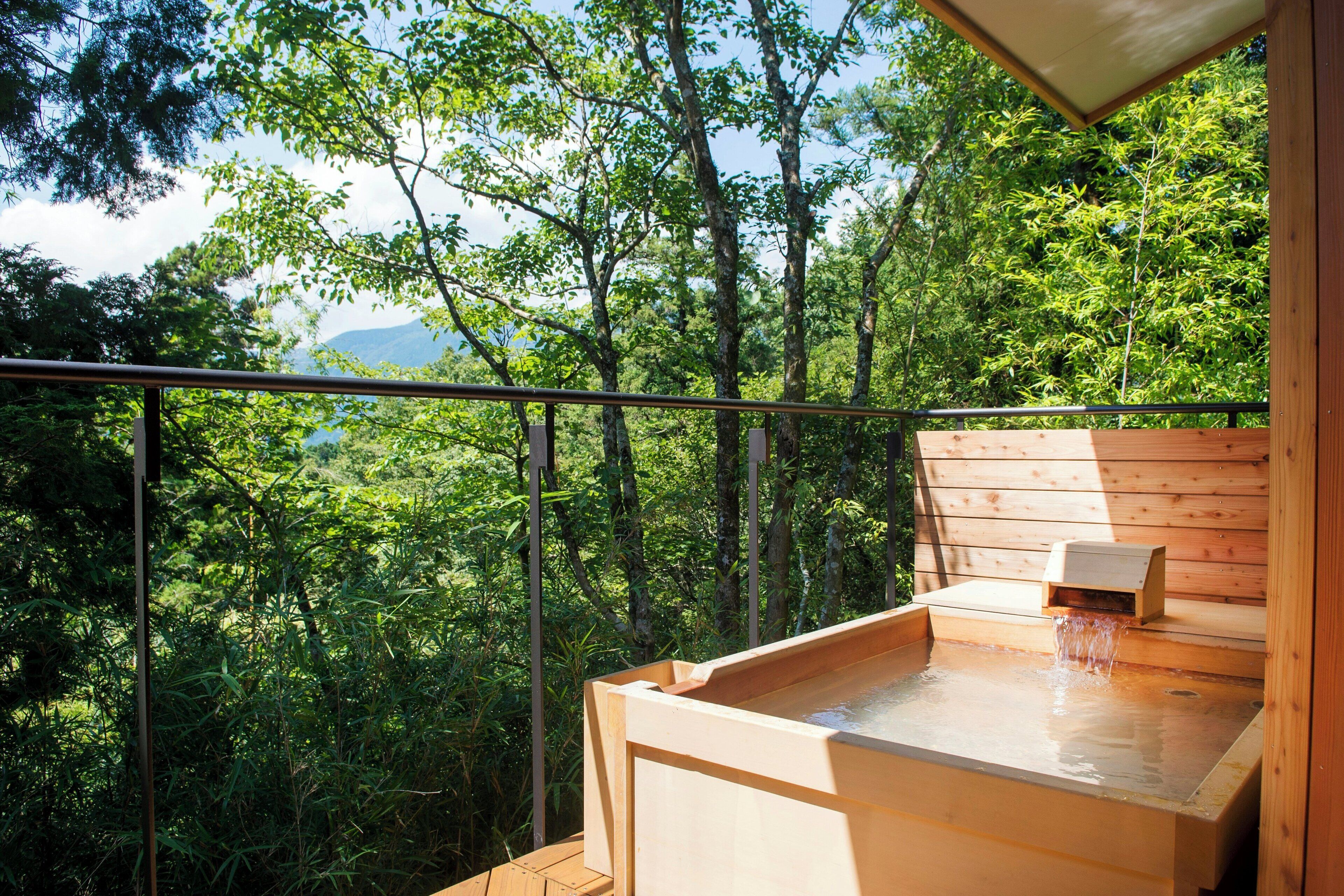 A wooden hot tub with a waterfall feature, surrounded by lush greenery and a wooden deck.