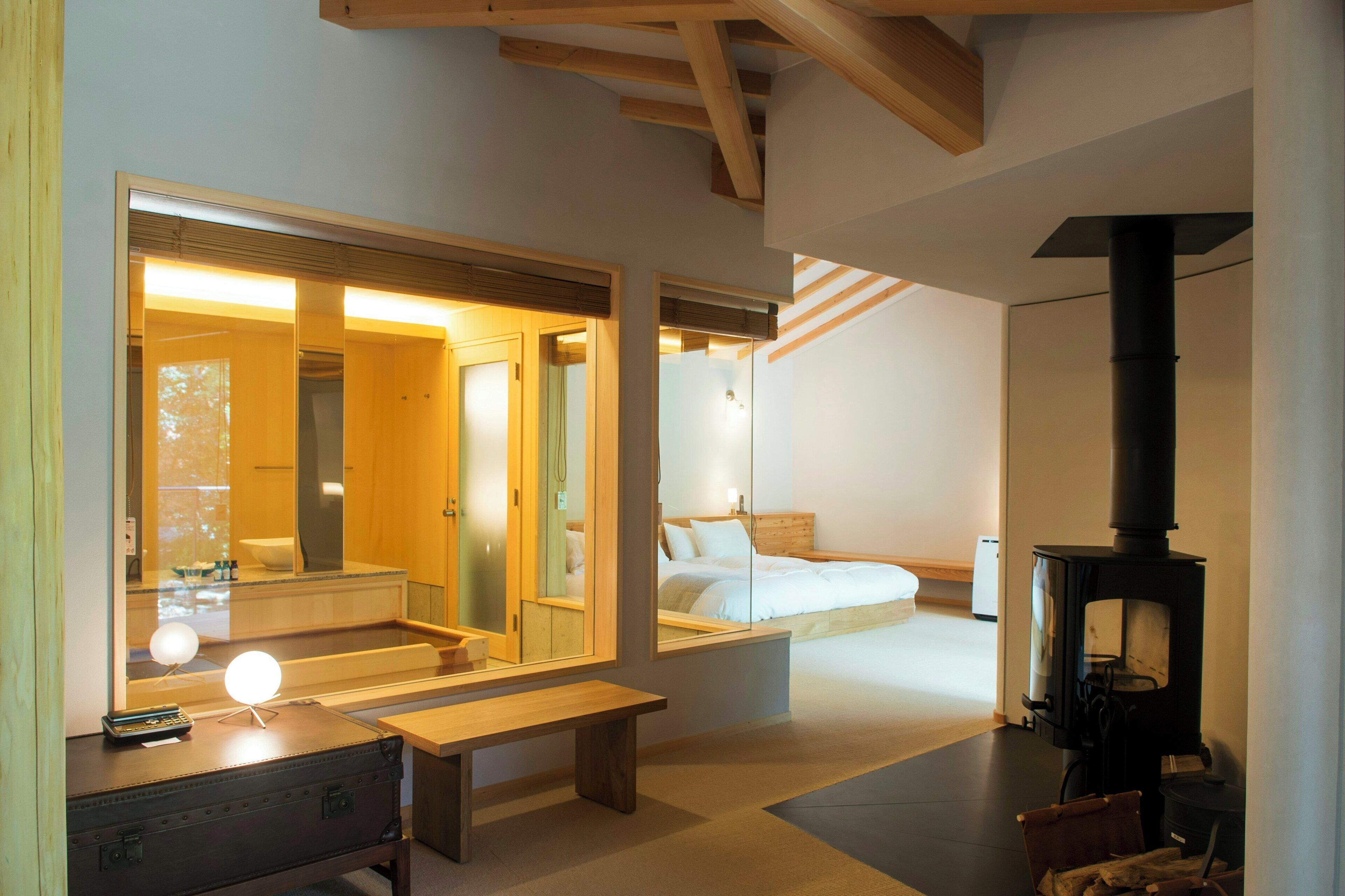 A modern bedroom with a wooden bench, a mirror, a bedside table with a lamp, and a black wood-burning stove.