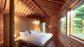A wooden cabin bedroom with a large bed, a nightstand, and a window with a view of the forest.
