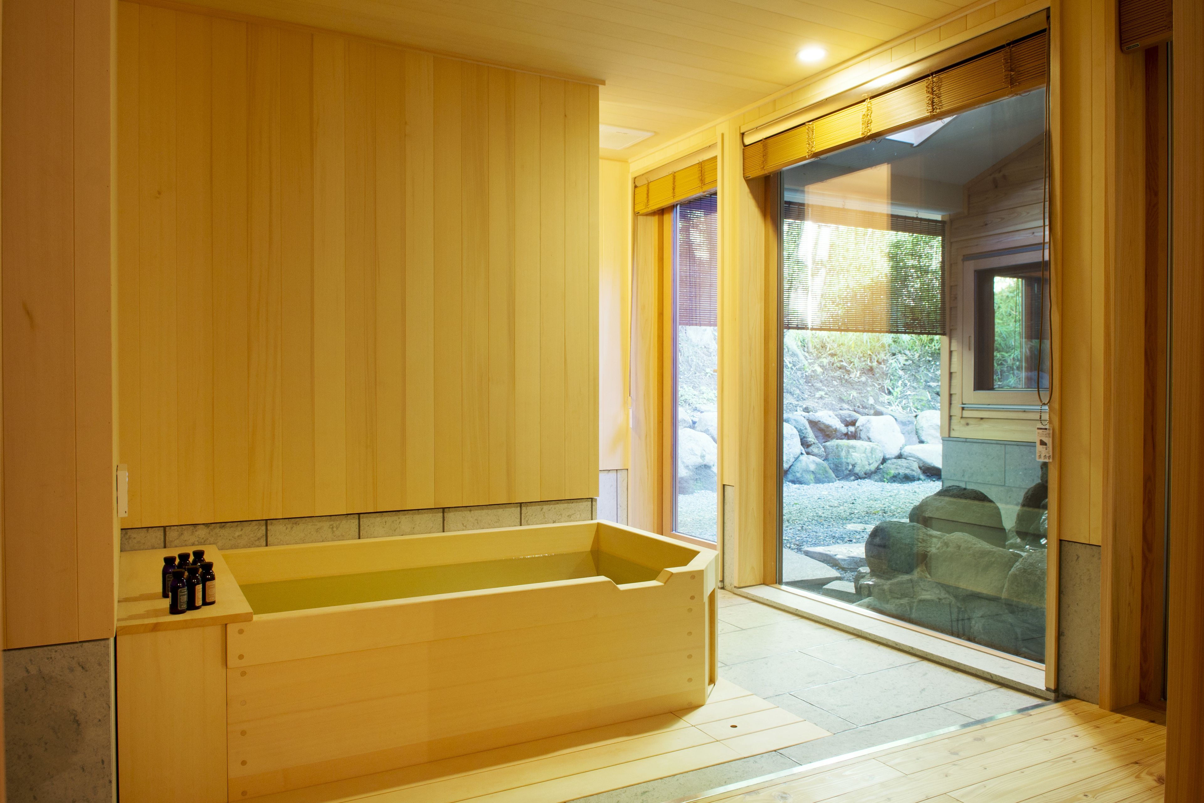 Ein Spa-Raum mit Holzboden, einer großen Badewanne, Holzschränken und einer Glastür, die zu einem Außenbereich mit Steinen führt.