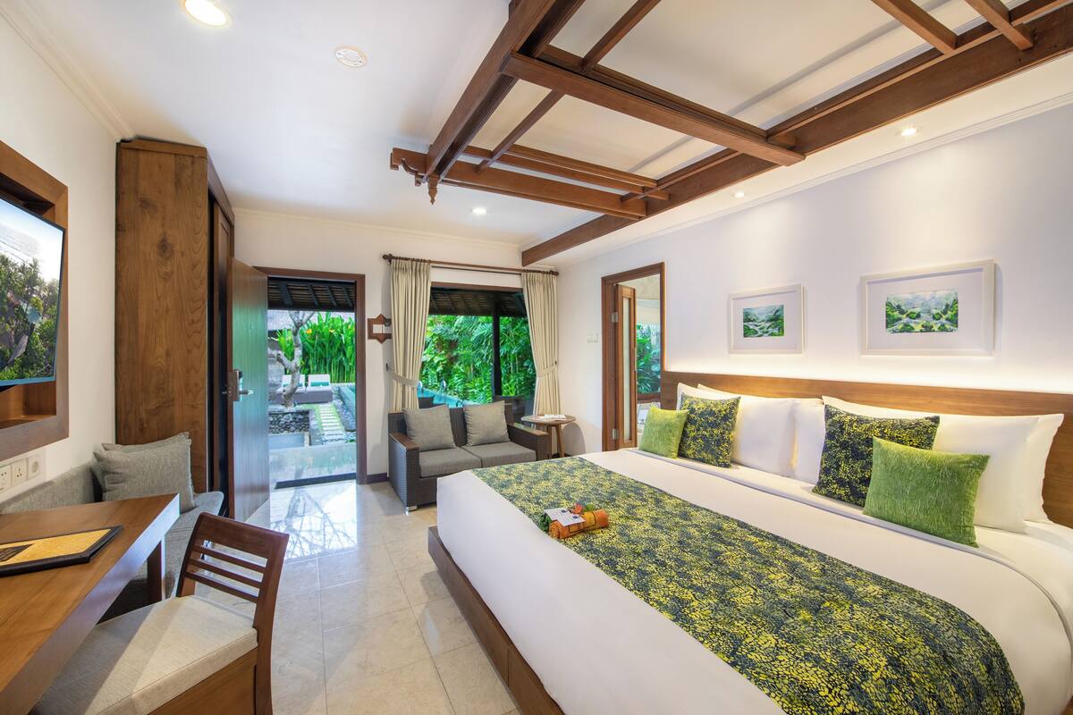 A hotel room with a large bed, a dining area, and a view of greenery outside.