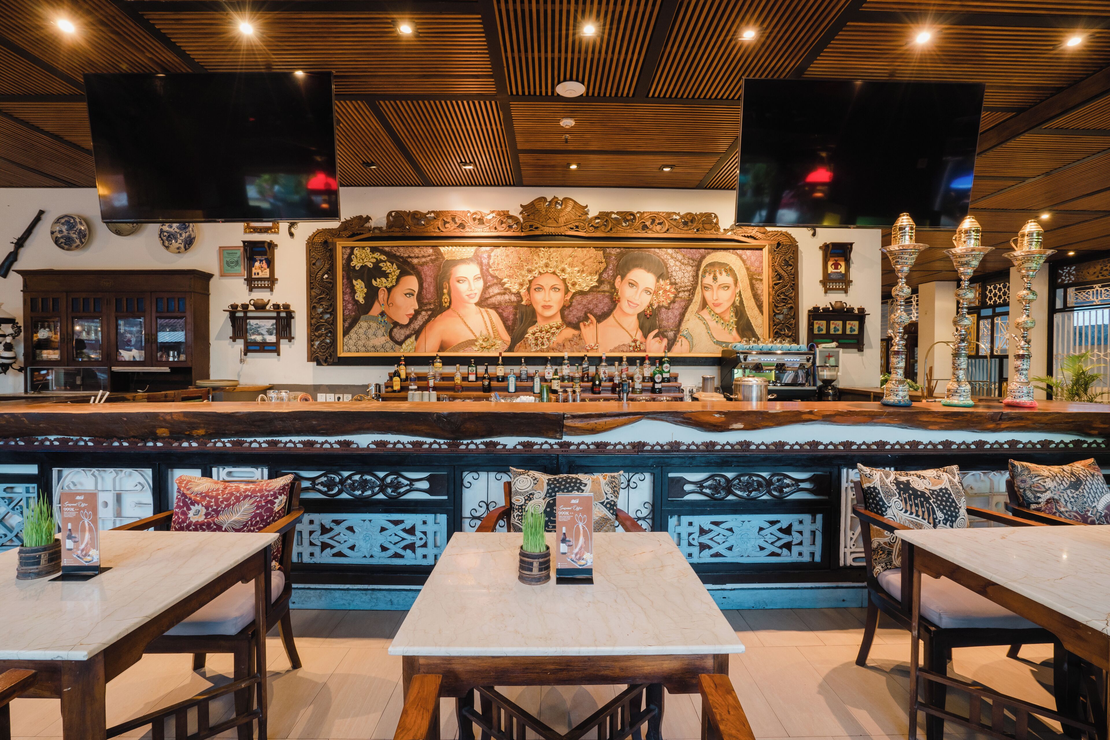 A bar area with a wooden ceiling, a large mural, and a bar counter with bottles. There are tables and chairs in the foreground.