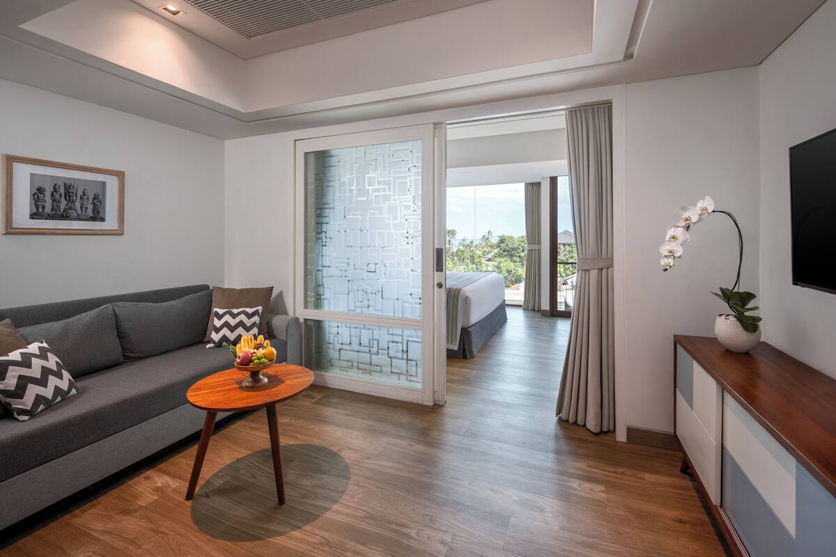Une chambre d’hôtel moderne avec un canapé, une petite table en bois sur laquelle sont disposés des fruits, une porte vitrée donnant sur un balcon et un téléviseur fixé au mur.