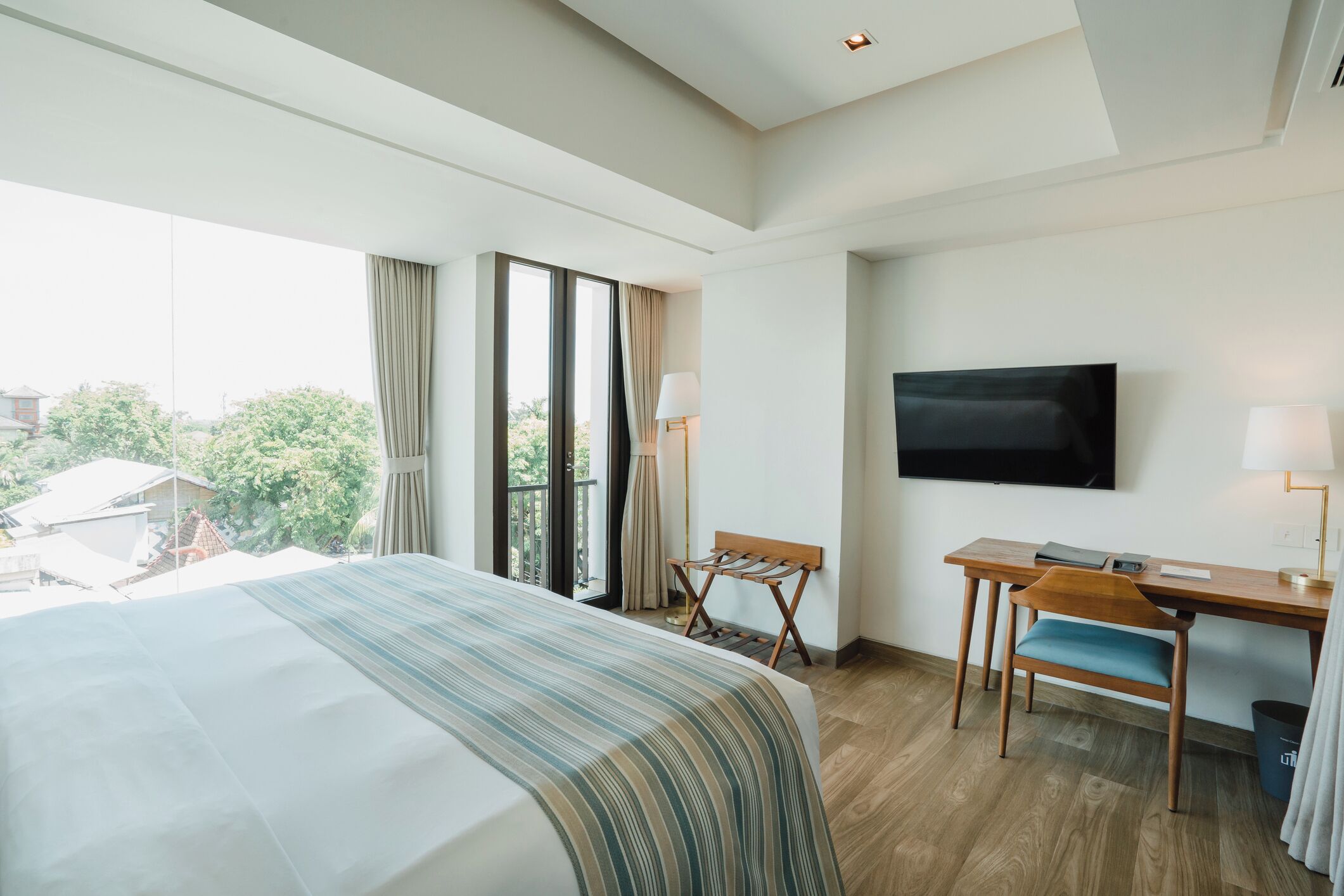 A hotel room with a large bed, a desk with a chair, a flat-screen TV mounted on the wall, and a balcony with curtains.