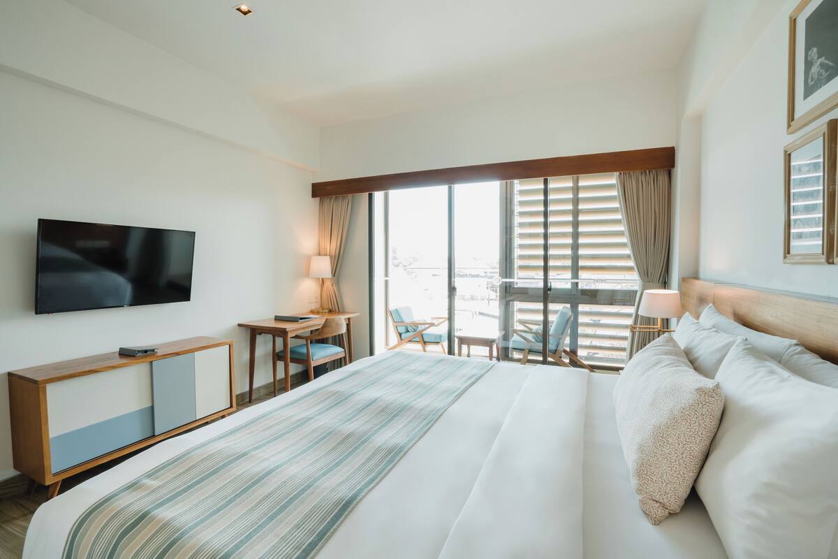 Une chambre d’hôtel dotée d’un grand lit, d’un téléviseur fixé au mur, d’un bureau avec une lampe et offrant une vue sur l’extérieur grâce à de grandes fenêtres.