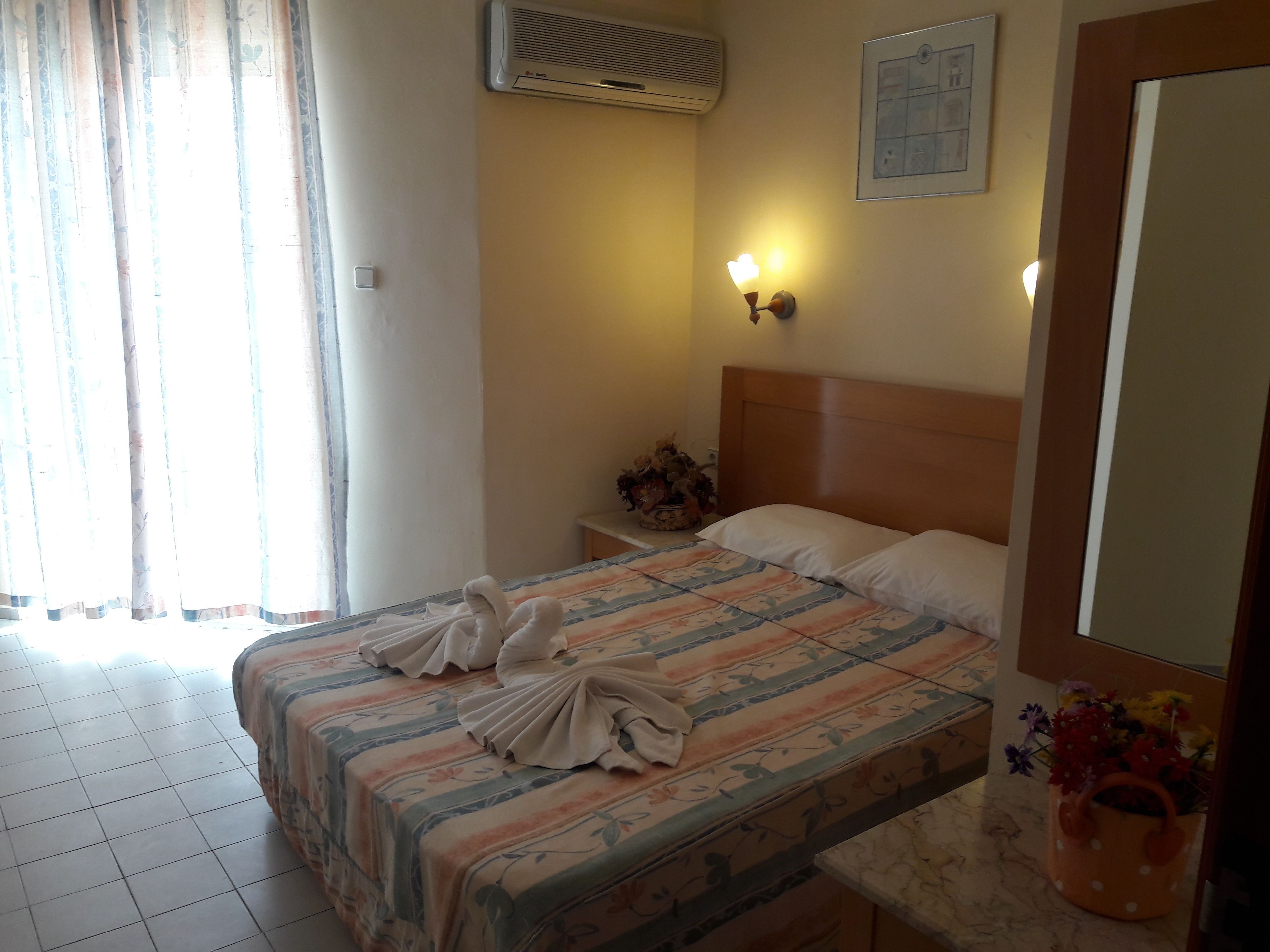 A hotel room with a bed, a nightstand, a wall-mounted air conditioner, a wall clock, and a window with curtains.
