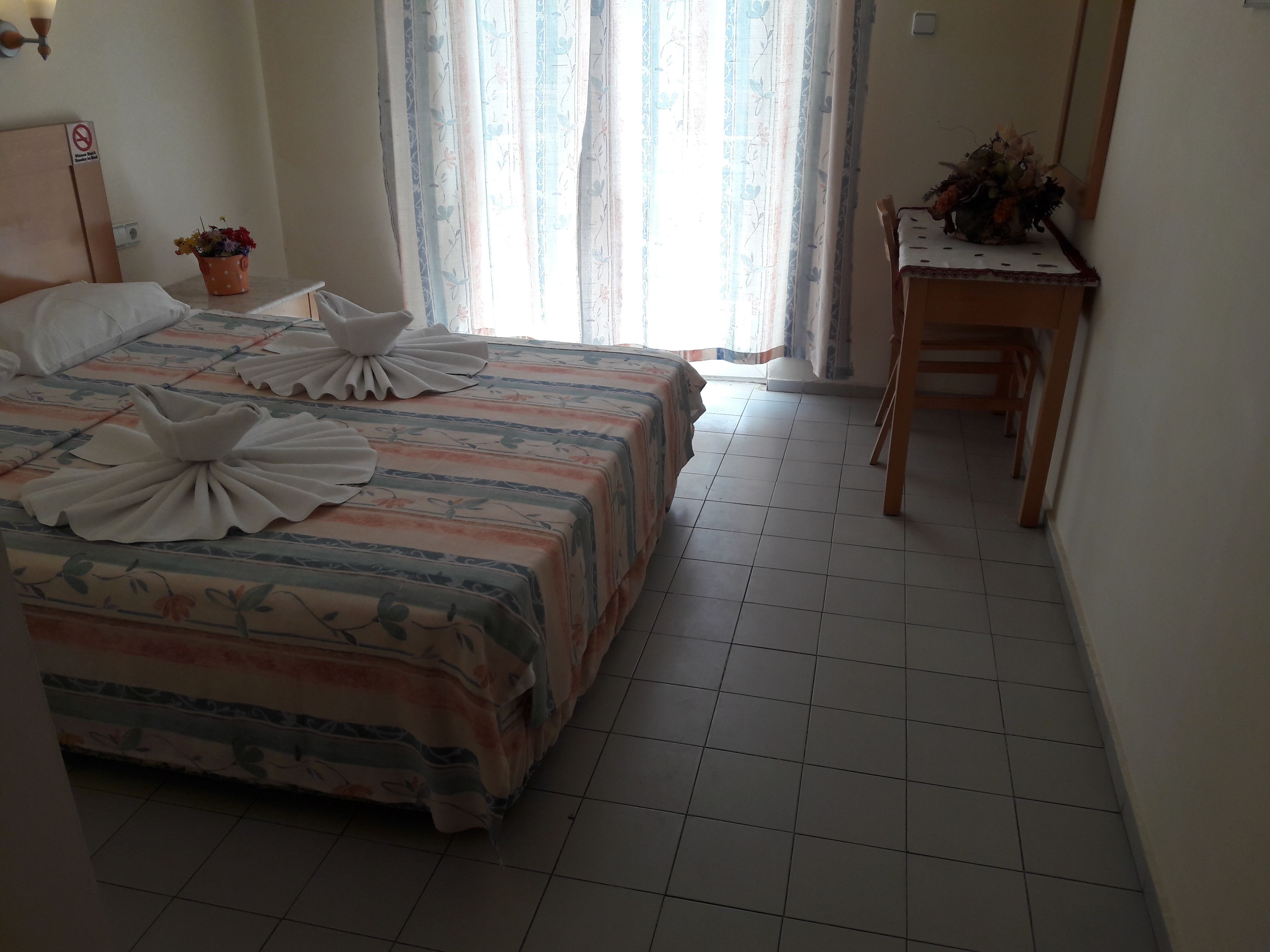 Una cama bien hecha con almohadas decorativas y una colcha estampada con motivos florales. Hay una mesita con un jarrón de flores y una silla.