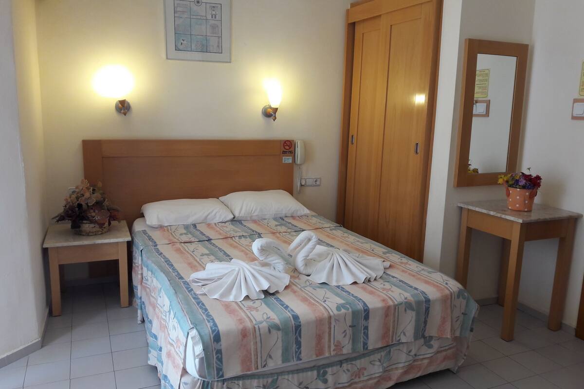A neatly made bed with a floral bedspread, two bedside lamps, a wooden headboard, a wooden wardrobe, a small bedside table with a vase, and a mirror on a wooden console table.