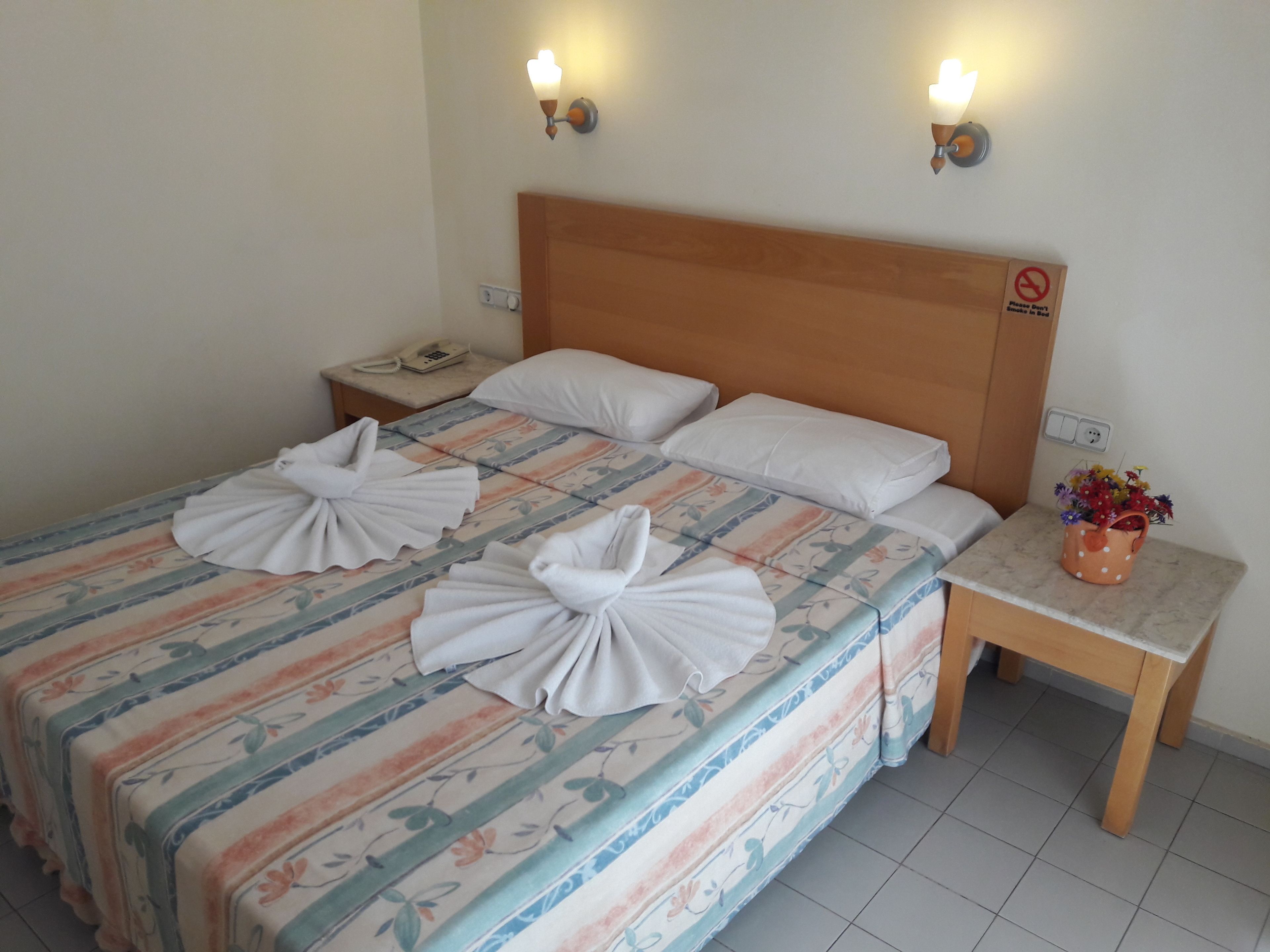 Une chambre d’hôtel comprenant un lit, deux tables de chevet, une petite table, un téléphone et un vase à fleurs.