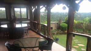 Un espacio al aire libre cubierto con una mesa redonda de vidrio y sillas, vista del paisaje circundante y una estructura de techo de madera.