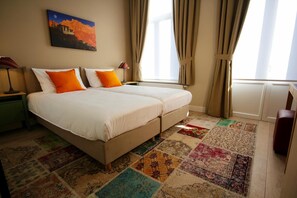 Una cama doble con dos almohadas, una mesita de noche, una lámpara, una ventana con cortinas y una alfombra colorida.
