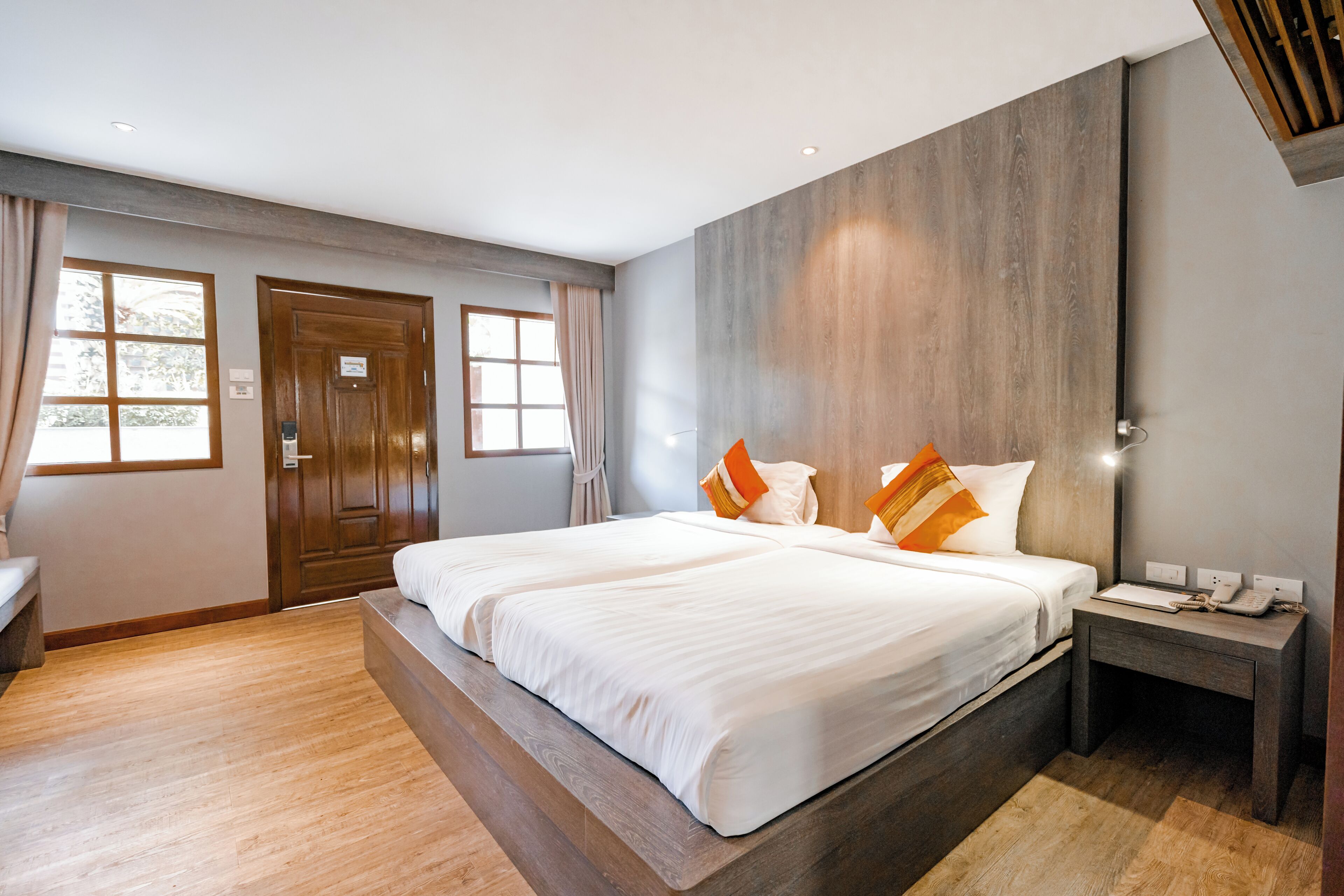 A double bed with white linens and orange pillows, a wooden nightstand with a phone, and a large window with curtains.