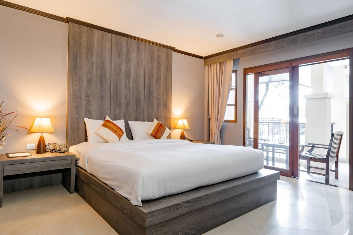 A large bed with a wooden headboard, two bedside lamps, a nightstand, and a view of the outside balcony.