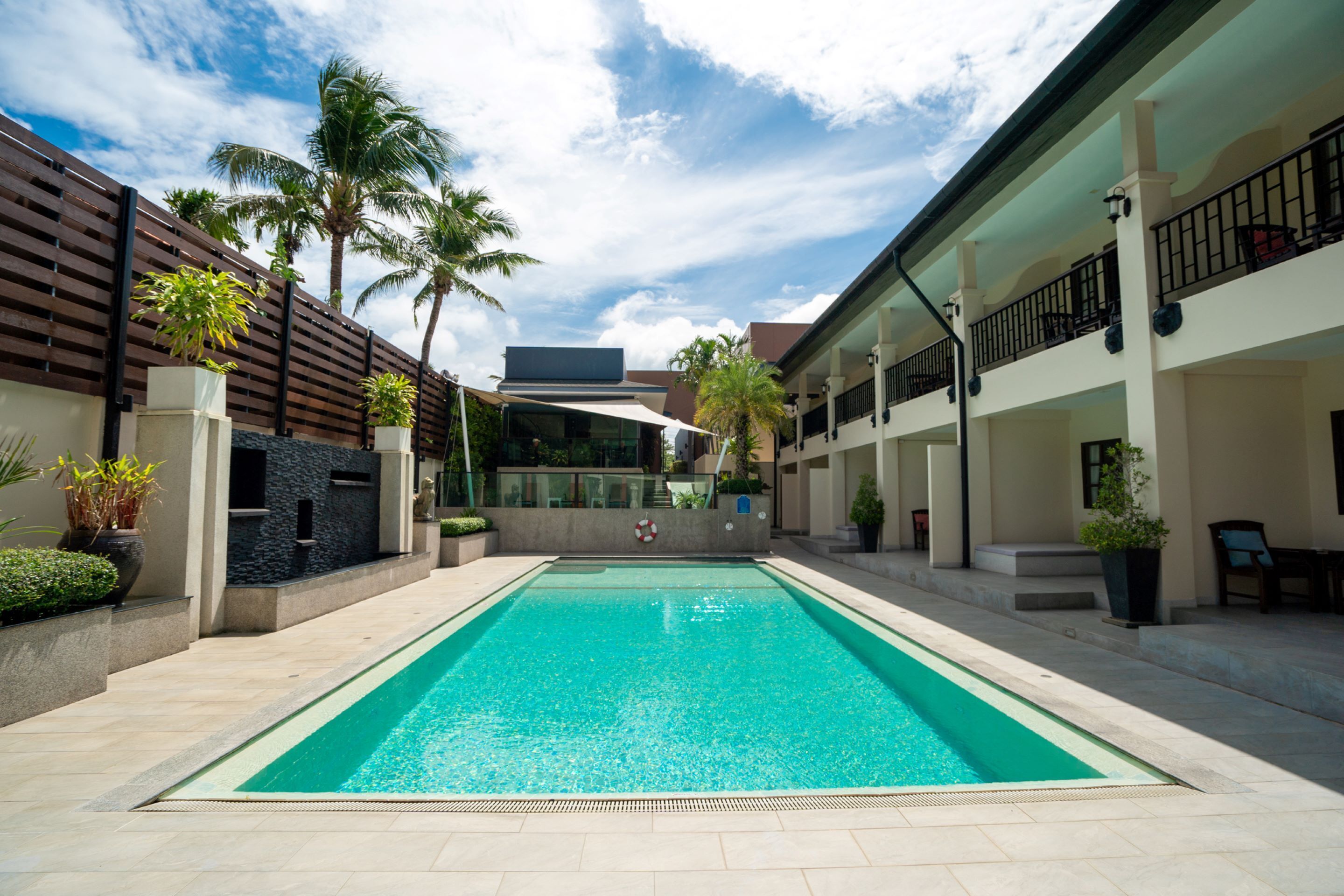 A rectangular swimming pool with a tiled deck, surrounded by a wooden fence and palm trees.