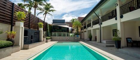 A rectangular swimming pool with a tiled deck, surrounded by a wooden fence and palm trees.