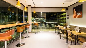 Een modern café met houten tafels en stoelen, gele hanglampen en uitzicht op groen door grote ramen.