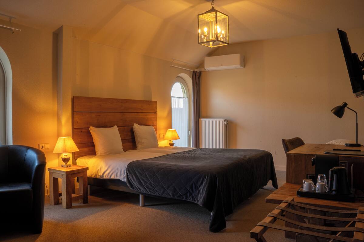 A hotel room with a bed, bedside lamps, a wooden headboard, a small table with a lamp, a chair, a desk with a coffee maker, and a television.
