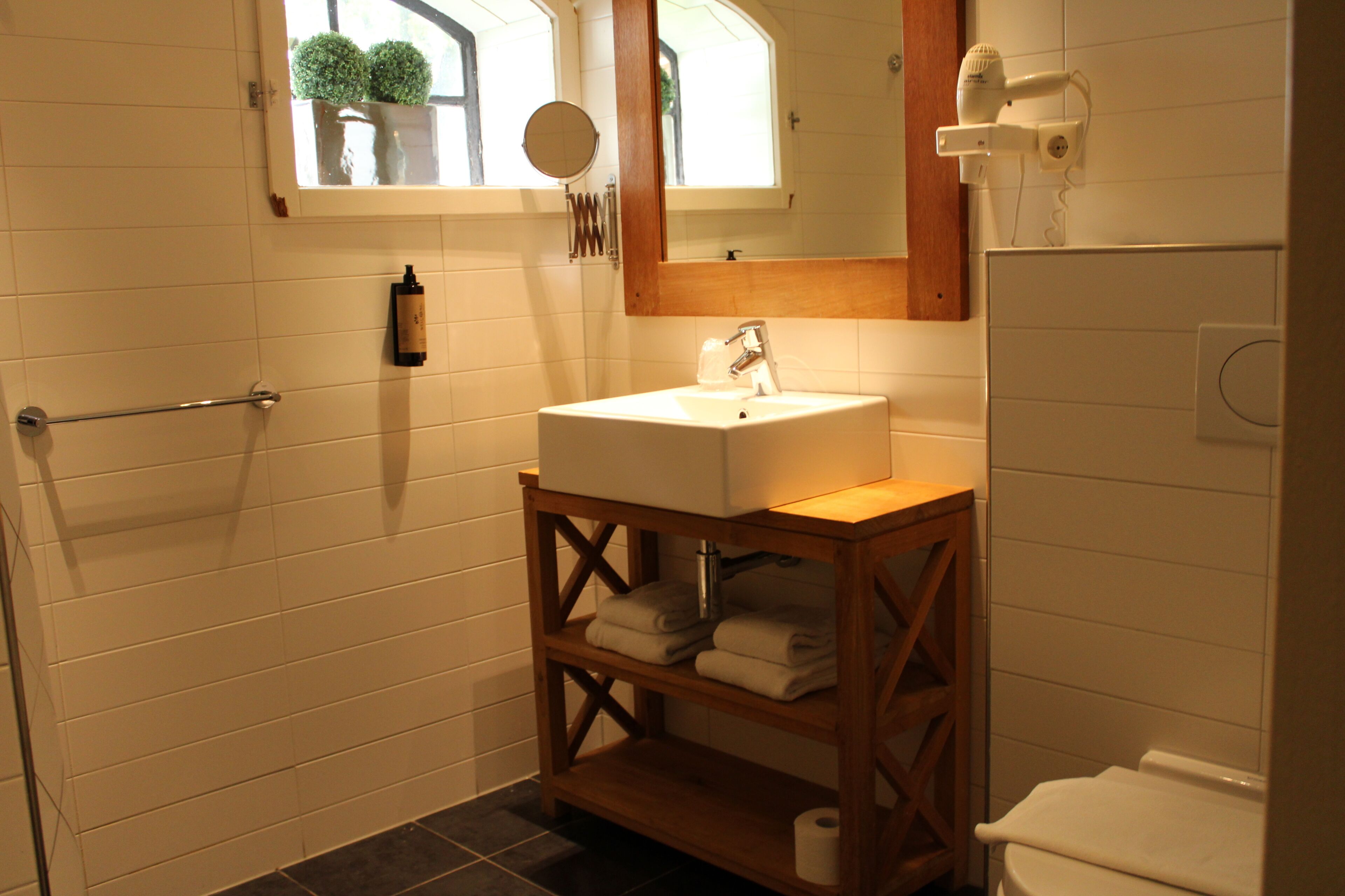 Un bagno con lavandino bianco, mensole in legno, porta asciugamani, uno specchio e una finestra con piante.