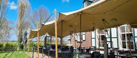An outdoor seating area with tables and chairs under a canopy, surrounded by a brick building and trees.