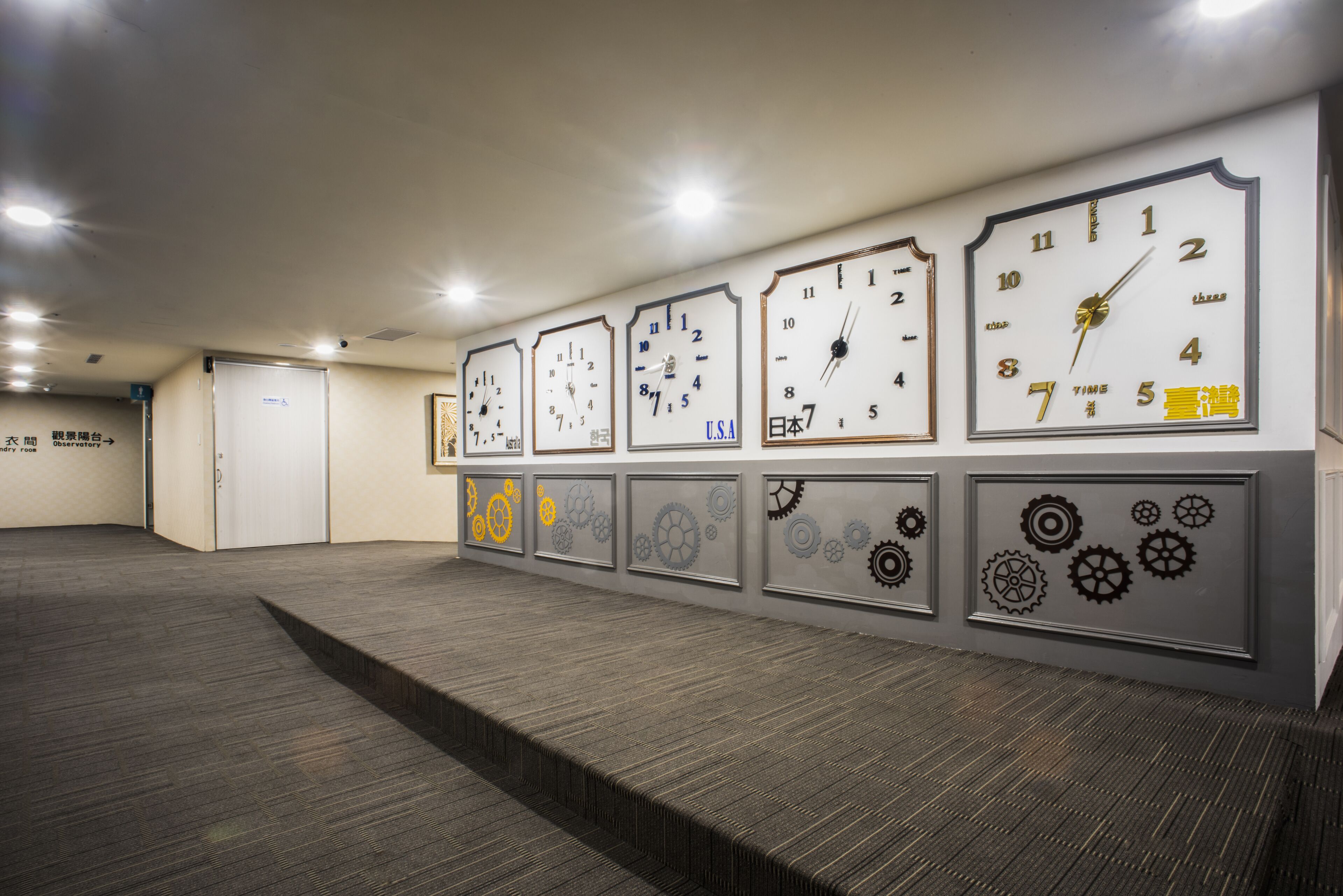 Un pasillo con varios relojes en la pared, suelo alfombrado y puertas a la izquierda.