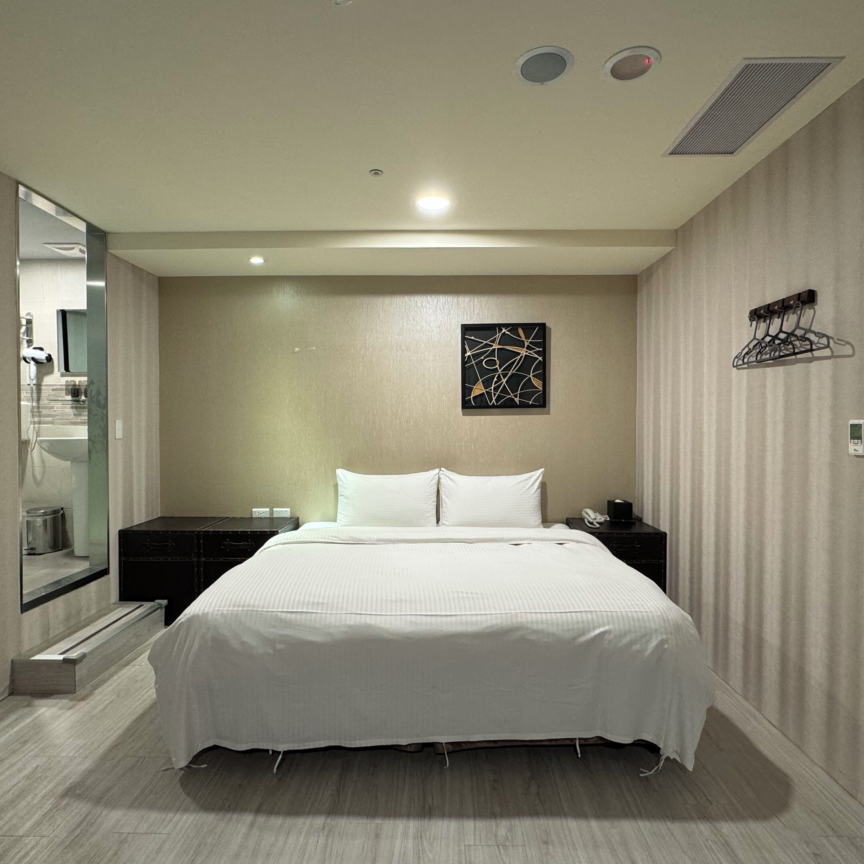 Habitación de hotel con cama, mesitas de noche, una obra de arte en la pared, perchero y baño visible a través de una puerta de cristal.