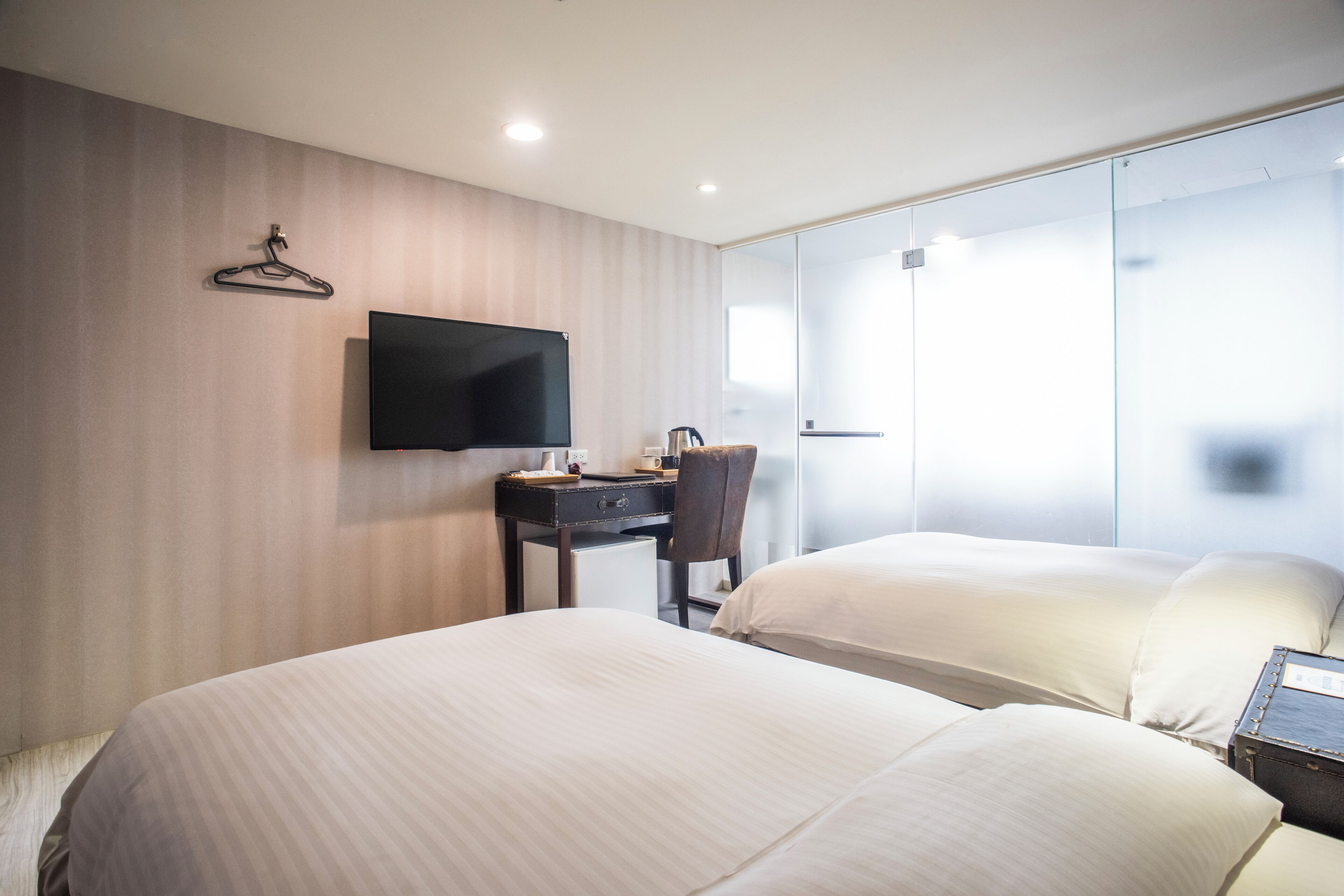A hotel room with two beds, a desk, a chair, a TV, and a large window.