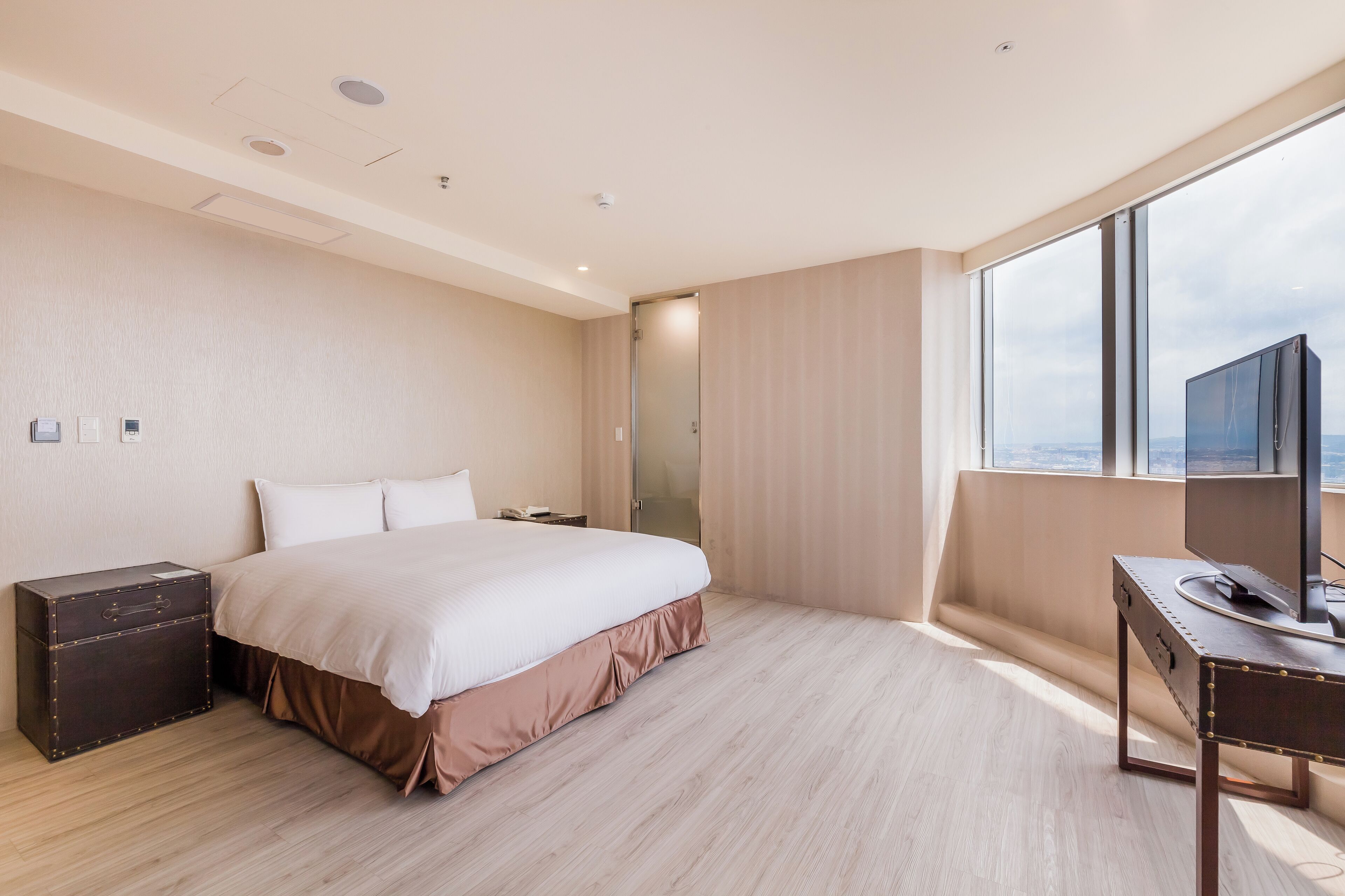 Habitación de hotel moderna con una cama grande, un escritorio con una computadora, una maleta y vistas a la ciudad a través de amplias ventanas.