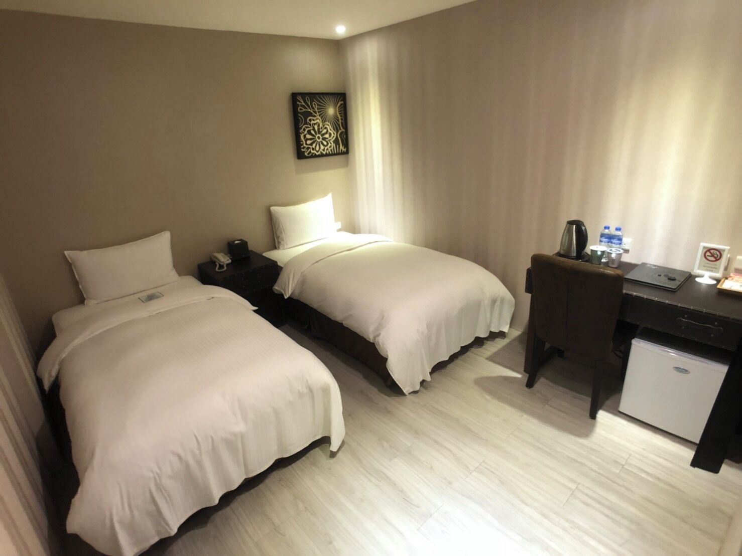 A hotel room with two beds, a desk, a chair, a small fridge, and a decorative wall piece.